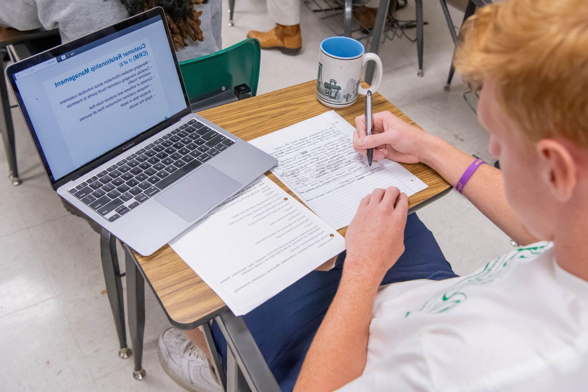 坐在教室课桌前的学生, taking notes on paper, 看着笔记本电脑上打开的ppt.